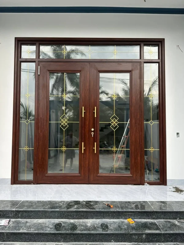 Cửa chính vân gỗ nâu đỏ, lan đồng hình thoi lồng nhau, phía trước là bậc đá đen