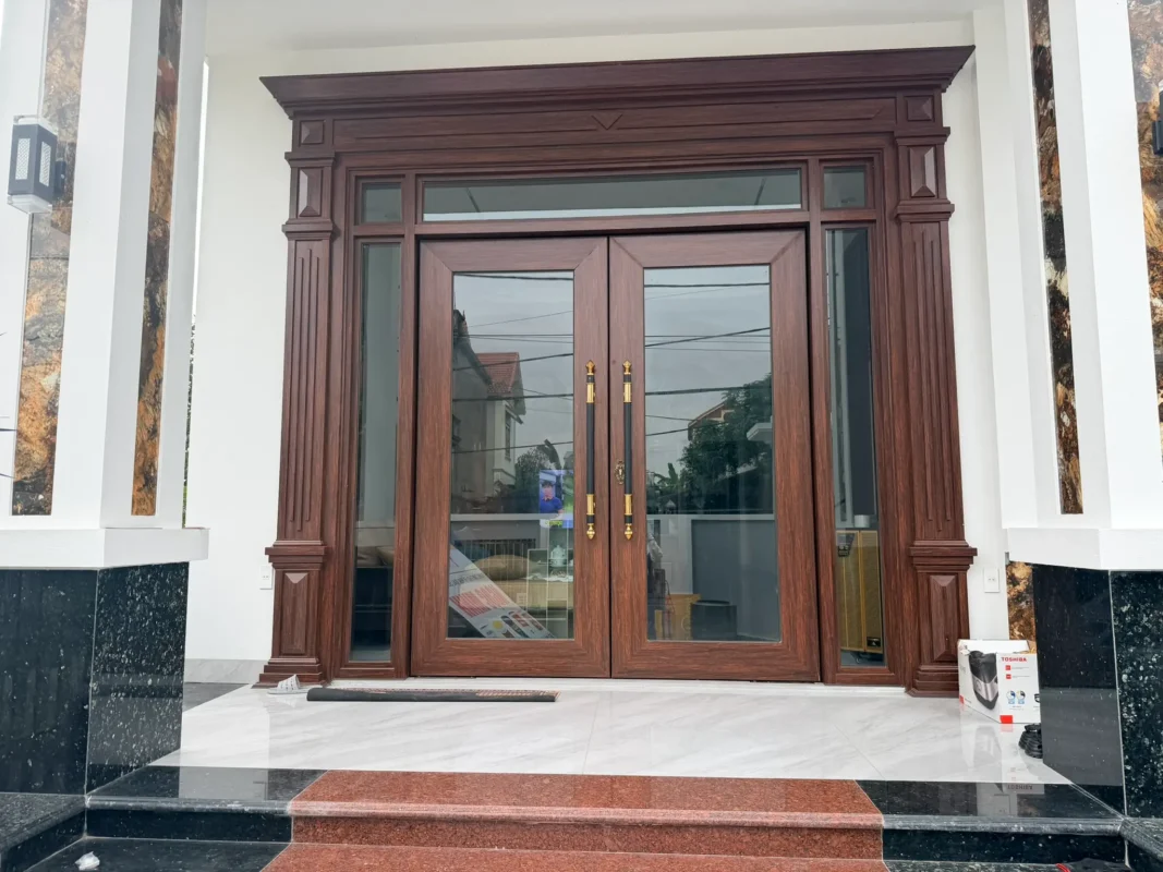 Cửa chính vân gỗ có phào đại phía trên, hai bên là cột ốp đá