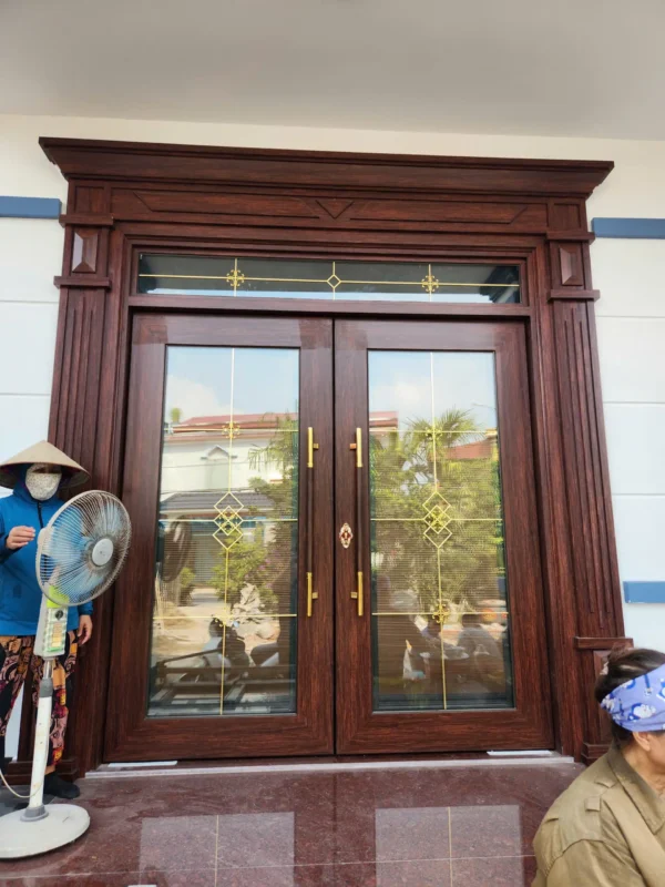 Cửa đi hai cánh nhôm vân gỗ lắp trong khung phào chỉ, có ô kính thoáng phía trên cửa