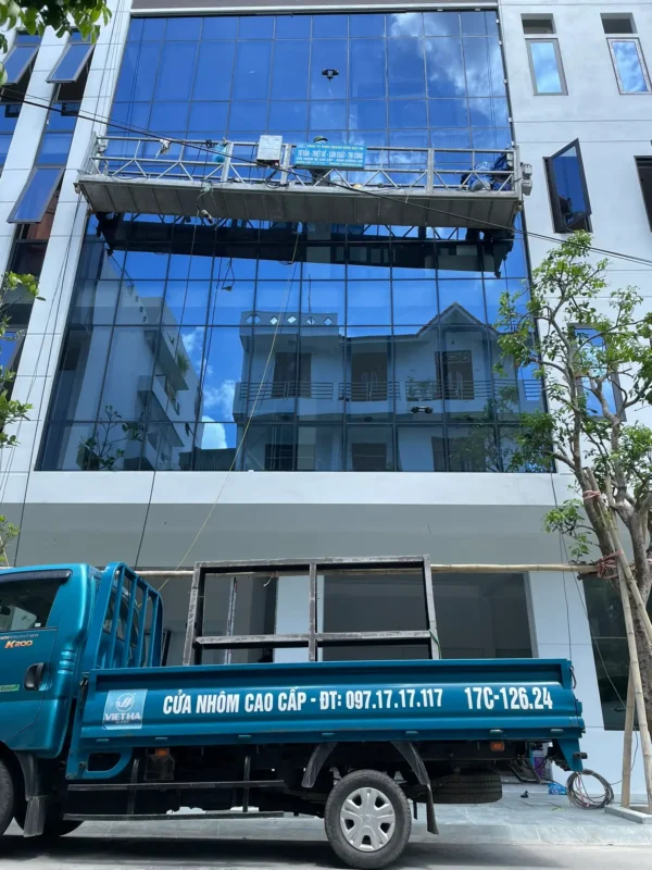 Xe tải Nhôm Kính Việt Hà chở giá đỡ kính, đỗ trước mặt dựng kính xanh đang lắp bằng gôn nâng hạ