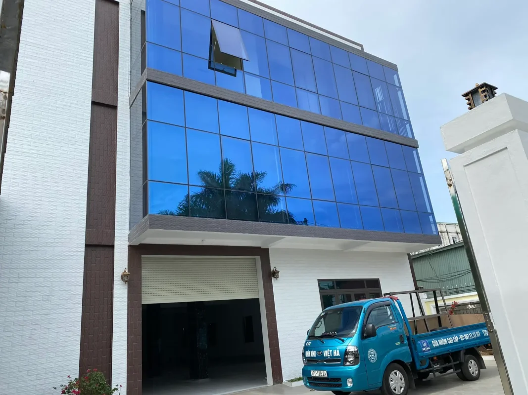 Mặt dựng kính màu xanh ba tầng có một ô cửa sổ mở hất, xe tải Nhôm Kính Việt Hà đỗ ở sân
