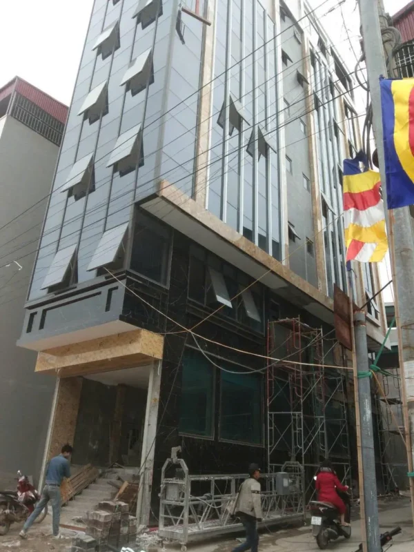 Tòa nhà mặt tiền ốp tấm màu xám và kính, cửa sổ mở hất, đang thi công với giàn giáo và giàn treo