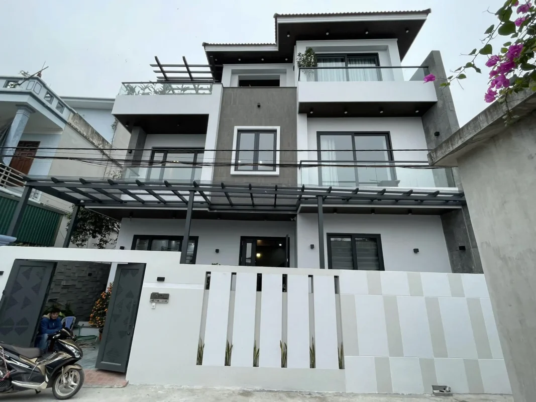 Nhà ba tầng có mái kính khung thép che tầng hai, lan can kính ban công và cửa sổ nhôm màu xám