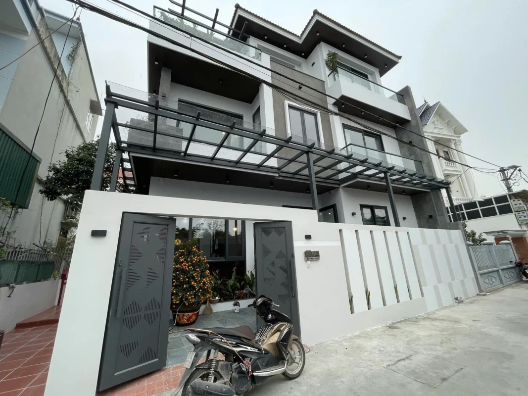 Mặt tiền nhà ba tầng nhìn từ cổng, mái kính khung thép, lan can kính và cửa nhôm màu xám