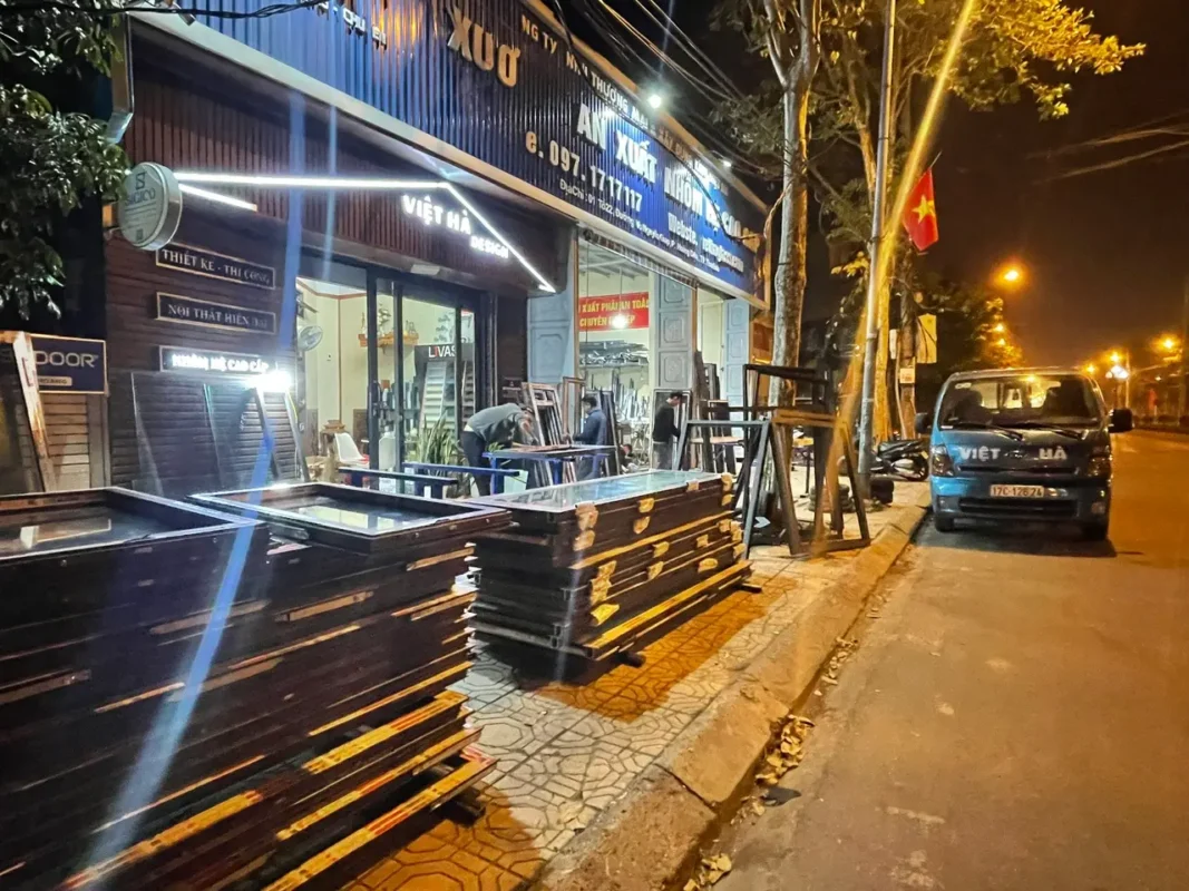 Thợ ghép khung cửa nhôm trên bàn làm việc trước xưởng Nhôm Kính Việt Hà, cánh cửa xếp chồng trên vỉa hè